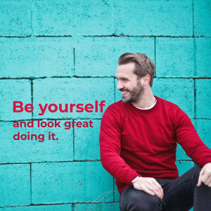 Man in red sweater smiling against a teal brick wall with motivational text.