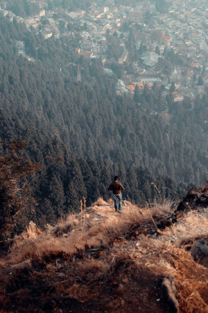 A lone figure stands on a cliff overlooking a vast valley and forested mountains.