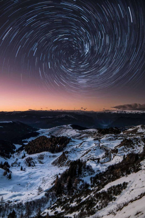 Long exposure photograph of star trails circling over a snow-covered mountain landscape.