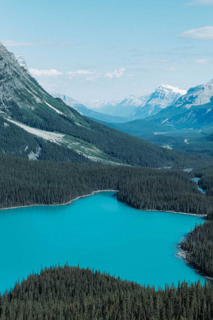 Aerial view of a vibrant turquoise lake surrounded by evergreen forests and snow-capped mountains.