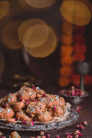 A close-up of a silver platter piled with Gulab Jamun, garnished with silver leaf and rose petals, with a blurred festive background.