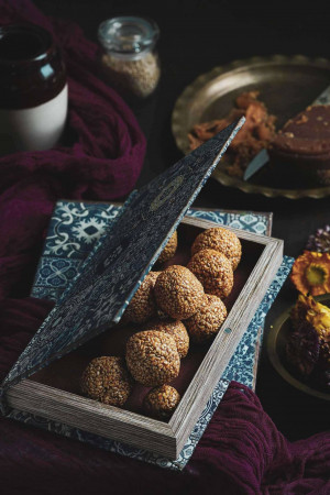 Close-up of sesame seed sweets in a wooden box with a blue patterned lid, surrounded by traditional Indian sweets and fabrics.