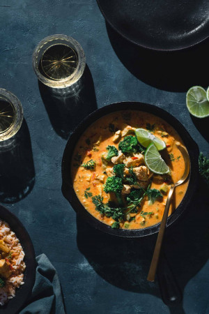 Overhead shot of a vibrant Thai red curry served in a black bowl with lime wedges and a glass of drink.