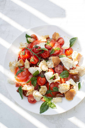 Overhead shot of a vibrant tomato and mozzarella salad with croutons and basil on a white plate.