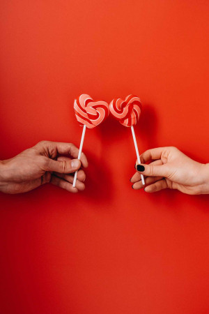 Two hands holding red swirl lollipops in the shape of hearts against a red background.