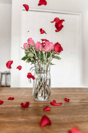 A glass vase with pink and red roses on a wooden table, with rose petals falling around it.