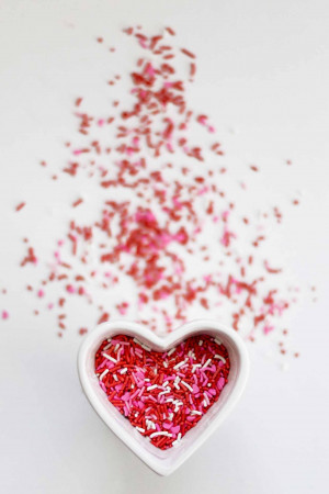 A white heart-shaped bowl filled with red, white, and pink sprinkles on a white background.