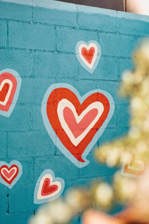 A colorful mural of red hearts on a teal brick wall with blurred flowers in the foreground.