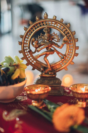 Golden Nataraja statue surrounded by fruit, flowers, and lit oil lamps.