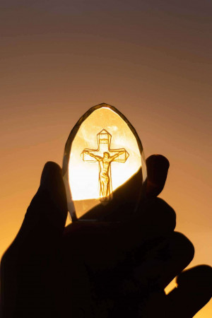 A hand holding a crystal with a crucifix visible inside, backlit by a sunset.