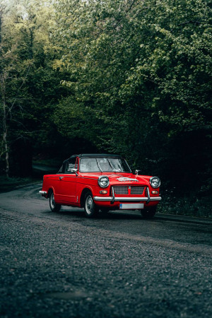 A red vintage Triumph sports car driving on a winding road through a forest.