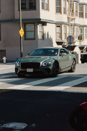 A green Bentley coupe parked on a street with a crosswalk and buildings in the background.