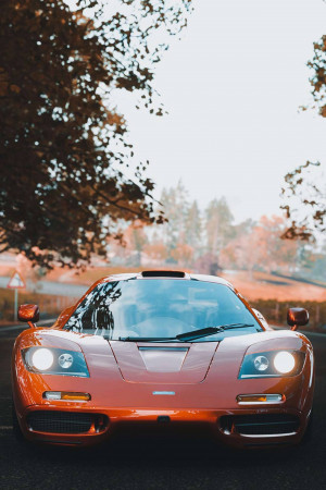 Front view of an orange McLaren F1 sports car on a road with autumn foliage in the background.
