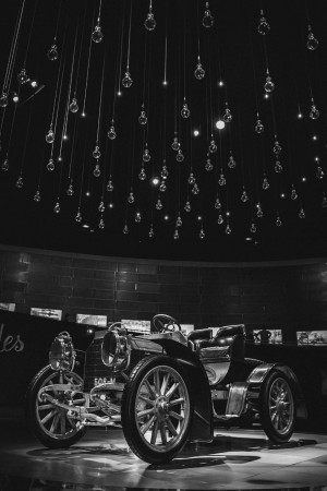 Black and white image of a classic car displayed under a ceiling of hanging light bulbs.