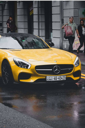 A bright yellow Mercedes-Benz parked on a wet city street with pedestrians walking by.