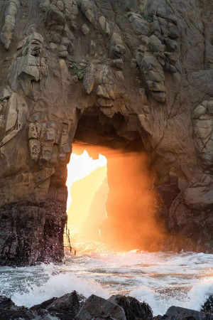 A natural rock archway on a coastline with waves crashing and a sunset visible through the opening.