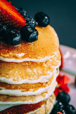 Close-up of a stack of golden pancakes topped with fresh strawberries and blueberries, drizzled with syrup.