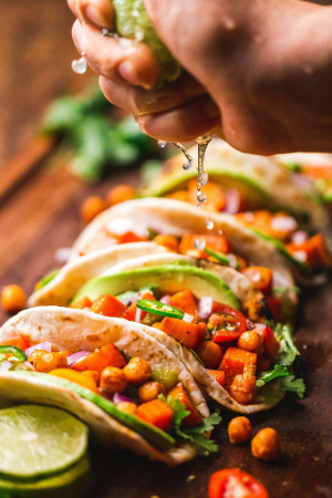 Close-up of hands squeezing lime juice over sweet potato tacos on a wooden surface.