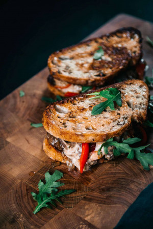Close-up of a toasted sandwich with mushrooms, peppers, and arugula on a wooden board.