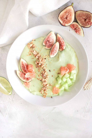A white bowl filled with a vibrant green smoothie, topped with fresh figs, grapefruit, cucumber, granola, and a lime wedge.