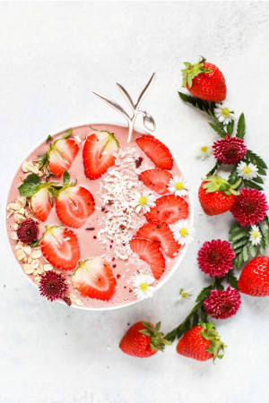 Top-down view of a pink strawberry smoothie bowl decorated with sliced strawberries, granola, coconut flakes, mint, flowers, and a golden fork.