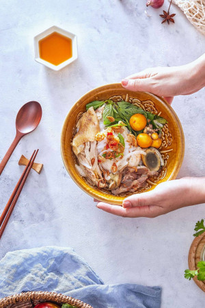 Overhead shot of a bowl of Vietnamese Pho with quail eggs, herbs, and meat, held by hands.