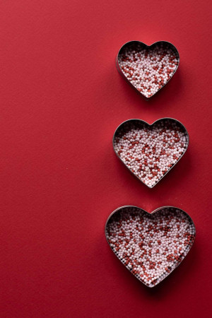 Overhead view of three heart-shaped metal cutters filled with white and red sprinkles on a red background.