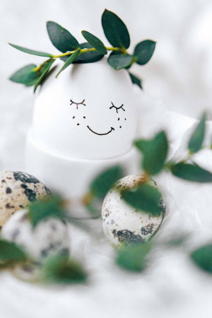 A white egg with a drawn smiling face and a eucalyptus crown, surrounded by other decorated eggs.