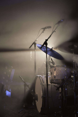 A dimly lit stage with a drum set and microphones, bathed in spotlights and smoke.