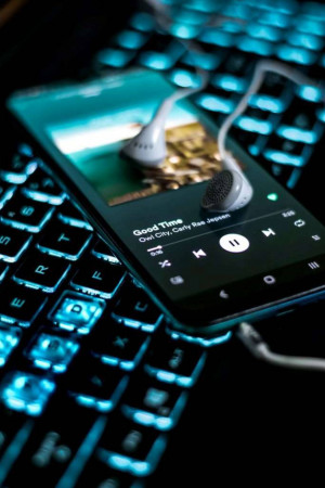 Close-up of a smartphone displaying a music streaming app interface with white earphones on top of a backlit keyboard.