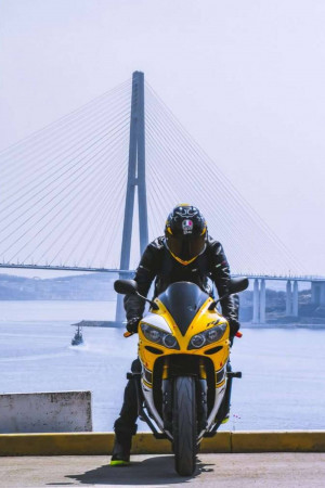 A motorcyclist in black and yellow gear riding a yellow motorcycle with a large bridge in the background.