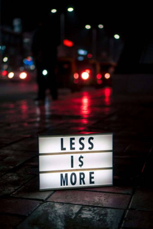 A lit sign reading 'LESS IS $' on a wet city street with blurred car lights in the background.