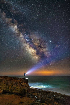 Silhouette of a person with a flashlight under a vibrant Milky Way galaxy over the ocean.