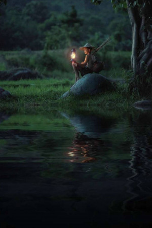 A young boy sitting on a rock by a river at night, holding a lantern.