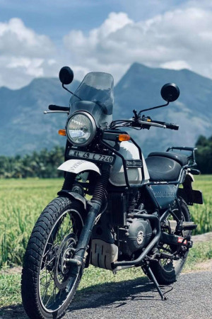 A silver Royal Enfield Himalayan motorcycle parked on a road with mountains in the background.