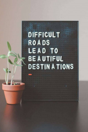 Black sign with white letters reading 'Difficult Roads Often Lead to Beautiful Destinations' with a small plant in a terracotta pot.
