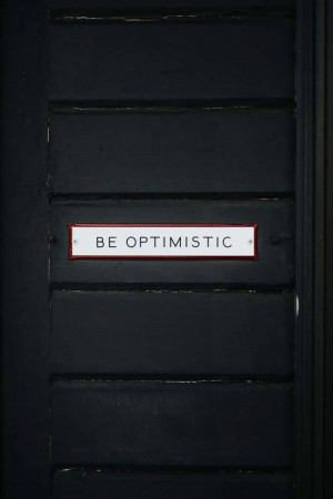 Close-up of a black wooden door with a red and white sign that reads 'BE OPTIMISTIC'.