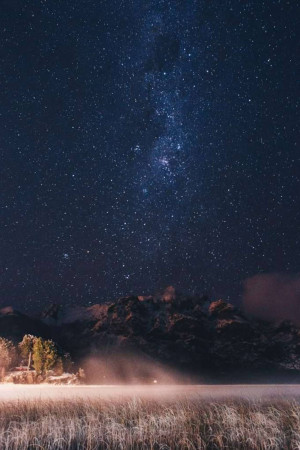 Night sky with the Milky Way galaxy over a silhouetted mountain range and illuminated field.