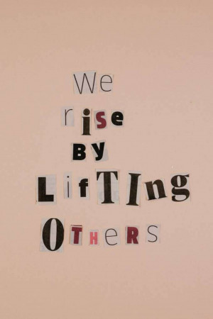 Cut-out letters spelling 'We Rise By Lifting Others' against a peach wall.