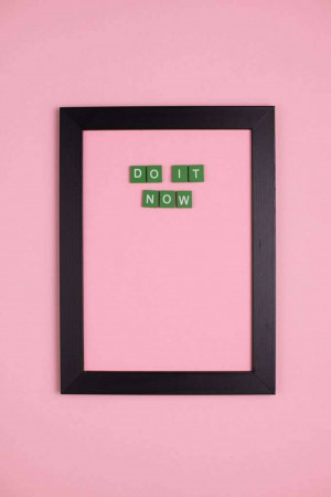 Black framed board with green letter tiles spelling 'DO IT NOW' against a pink wall.