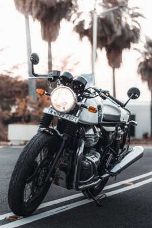 Silver and black Royal Enfield Continental GT motorcycle parked on a road with palm trees in the background.