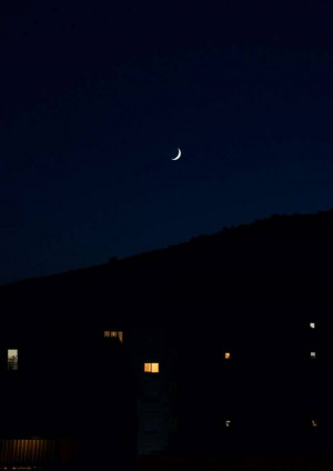 A crescent moon in a dark blue night sky over a town with illuminated windows.