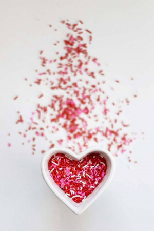 A white heart-shaped bowl filled with red and white sprinkles, surrounded by scattered sprinkles on a white surface.