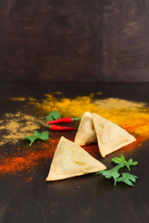 Close-up of three golden-brown samosas with red chili peppers and turmeric powder on a dark surface.