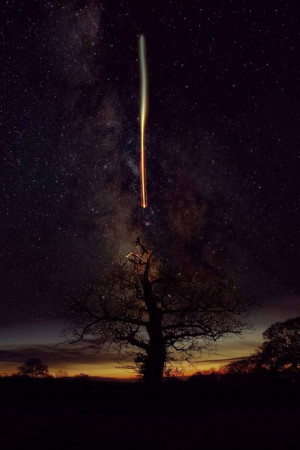 A long exposure photograph of the Milky Way with a bright shooting star over a silhouetted tree.