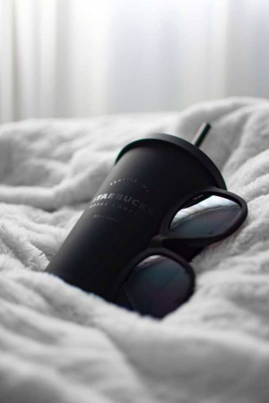 A black Starbucks reusable cup with a straw and a pair of sunglasses resting on a fluffy white blanket.