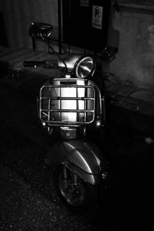 Black and white image of a classic Vespa scooter parked in a dimly lit area.