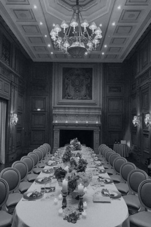 Long banquet table set for a formal dinner in a grand hall with a chandelier and fireplace.