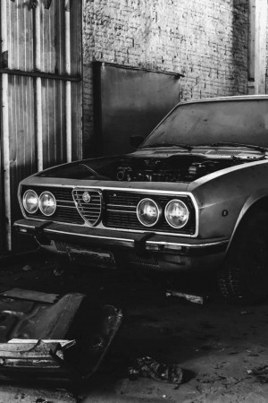 Black and white image of a classic Alfa Romeo car with the hood open, undergoing restoration in a garage.