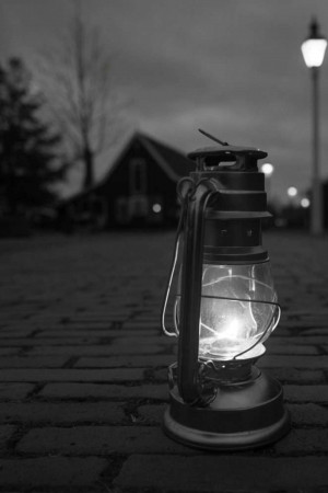 Close-up of an old-fashioned lantern lit against a dark, blurred background of a house and street lamp.